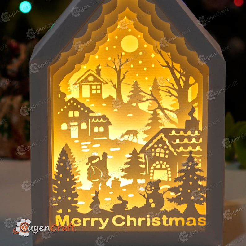 May include: A white wooden house-shaped light-up decoration with a cut-out design of a winter village scene. The scene includes a snowman, Santa Claus, a deer, trees, and houses. The words "Merry Christmas" are printed on the bottom of the house.