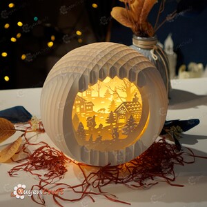 May include: A white paper sphere with a cut-out design of a winter village scene. The scene includes houses, trees, a snowman, and a person walking a dog. The sphere is illuminated from within, creating a warm glow.