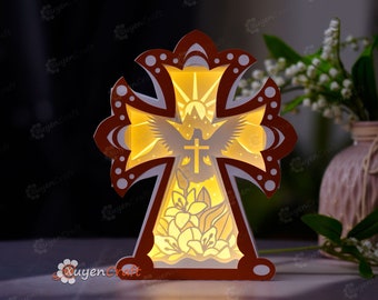 May include: A wooden cross-shaped light-up decoration with a dove and flowers inside. The cross is brown with white accents and the dove is white with a yellow halo. The flowers are yellow and white. The light is on and illuminates the design.