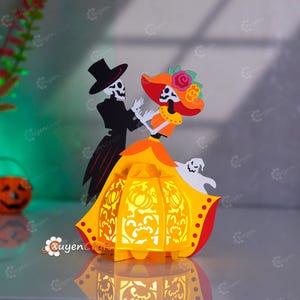 以下が含まれることがあります： フォーマルな衣装を着た踊る骸骨のカップルと幽霊が描かれた、紙製のランタン。ランタンは黄色とオレンジ色で、切り抜かれたデザインが施されています。骸骨は白と黒で、女性は大きな帽子をかぶっています。