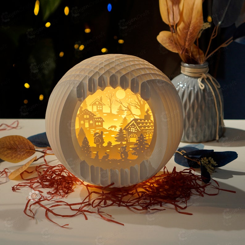 May include: A white paper lamp with a cut-out design of a winter village scene. The lamp is illuminated from within, casting a warm glow on the scene. The scene includes houses, trees, and a snowman.