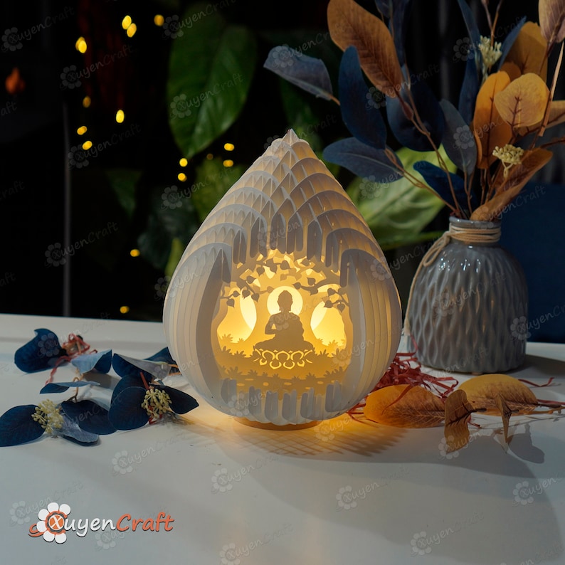 Op de afbeelding: Een witte lamp van papierkunst met een 3D-ontwerp van een mediterend Boeddhabeeld in een boom. De lamp is aan en geeft een warm licht.
