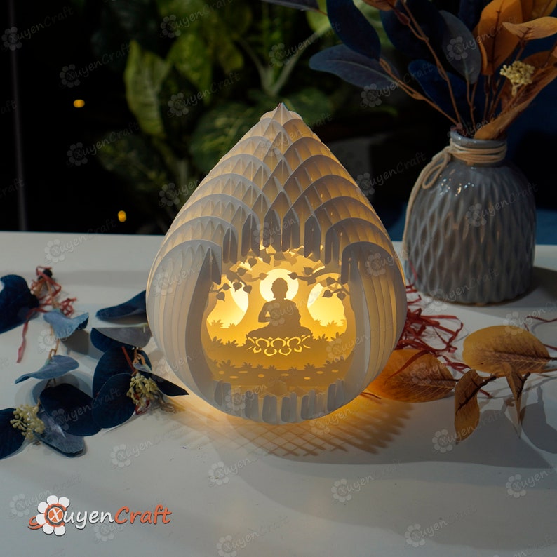 Op de afbeelding: Een witte papierkunstlamp in de vorm van een traan met een uitgesneden ontwerp van een mediterend Boeddhabeeldje erin. De lamp wordt van binnenuit verlicht en geeft een warm gloed.