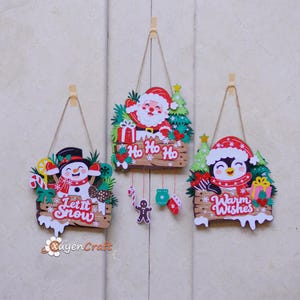 Könnte beinhalten: Drei hängende Weihnachtsschilder mit einem Schneemann und dem Text Let It Snow, dem Weihnachtsmann mit dem Text Ho Ho Ho und einem Pinguin mit dem Text Warm Wishes. Jedes Schild enthält bunte weihnachtliche Motive wie Geschenke und Tannen.