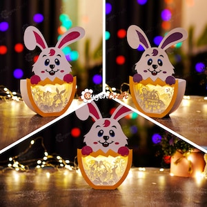 Puede incluir: Un conejo blanco asomándose de un huevo amarillo con una luz en su interior. El huevo tiene un diseño recortado de un conejo, flores y las palabras "Feliz Pascua".