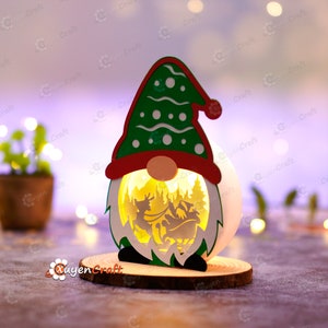 Pode incluir: Uma lanterna de Natal em forma de gnomo, com um chap&eacute;u verde e uma borda vermelha. A lanterna tem um corpo branco e redondo com uma cena recortada do Pai Natal e renas. Assenta numa base de madeira e &eacute; iluminada por dentro.