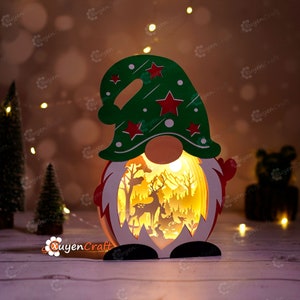 Peut inclure: Une figurine de gnome de Noël en bois avec un chapeau vert et une barbe rouge. Le gnome tient une scène illuminée d'une forêt d'hiver avec des cerfs et d'autres animaux.