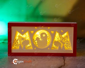 May include: A red wooden frame with a white interior, featuring a cutout design of the word "MOM" with a mother and child silhouette in the center. The frame is illuminated with warm white LED lights.