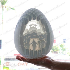 May include: White papercraft Easter egg with a 3D design of a cave with a cross and a figure of Jesus inside. The egg is being held in a hand.