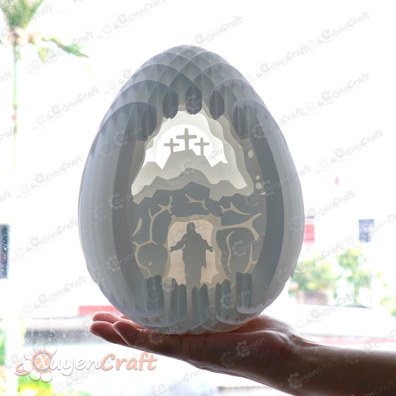 May include: White papercraft Easter egg with a 3D design of a cave with a cross and a figure of Jesus inside. The egg is being held in a hand.