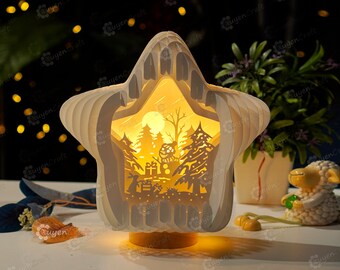 Könnte beinhalten: Eine weiße sternförmige Papierlampe mit einem ausgeschnittenen Design einer Winterszene. Die Lampe ist beleuchtet und strahlt ein warmes Licht aus. Die Szene zeigt einen Schneemann, Bäume und einen Mond.