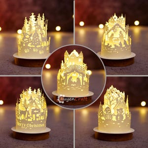 Puede incluir: Un conjunto de cinco lámparas cilíndricas blancas de papel recortado con iluminación interna cálida, cada una con escenas festivas intrincadas que incluyen un nacimiento, animales del bosque, muñecos de nieve y textos sobre bases de madera.