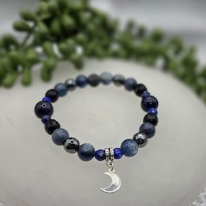 May include: A beaded bracelet featuring a mix of blue, black, and gray beads. A silver-colored crescent moon charm hangs from the bracelet. The beads vary in size and texture, creating a unique accessory.