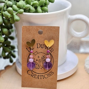 May include: A pair of purple and pink gemstone earrings with gold heart charms. The earrings are displayed on a brown card with the text "Lil Dipper Creations".