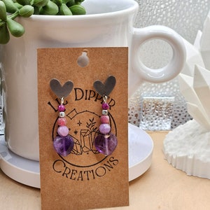 May include: A pair of purple and pink gemstone earrings with silver heart charms. The earrings are on a brown card with the text "Little Dipper Creations".