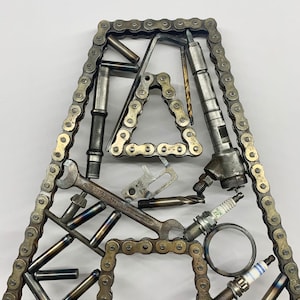 Könnte beinhalten: Eine Metallsculptur in Form des Buchstabens A, hergestellt aus einer Kette, Werkzeugen und anderen Metallteilen. Der Buchstabe besteht aus einer Kette und das Innere ist mit Werkzeugen wie Schraubenschlüsseln, Zündkerzen und anderen Metallteilen gefüllt.