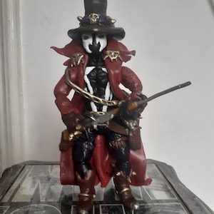 May include: A detailed action figure of a character in a red coat, black top hat, and white face paint. The figure has a chain around its neck and is holding a weapon. The figure is standing on a patterned surface.