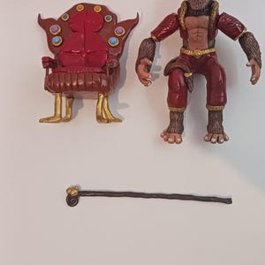 Puede incluir: Un juego de juguetes con una figura de mono, una silla con forma de trono y un bastón. La figura del mono es marrón y roja, con una cara detallada y texturas similares a la piel. La silla es roja con detalles dorados.
