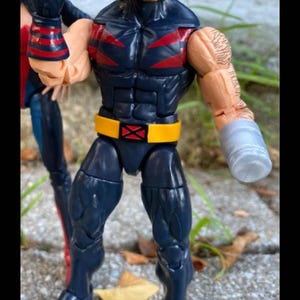 Può includere: Una action figure blu e rossa di Wolverine con accenti gialli e rossi. La figura ha artigli ed è in posa con un braccio alzato. La figura è in piedi su una superficie marrone con foglie.