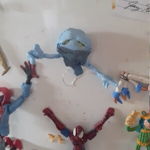 May include: A blue and gray clay figure with a creepy expression, hanging from a wire. The figure has long arms and legs. The figure is surrounded by other action figures, including a Spiderman figure with red and blue colors.