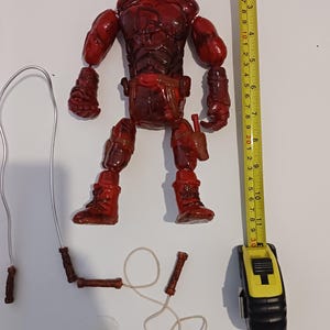 May include: A red and black action figure of Daredevil, a superhero with a red suit and a black mask, holding a pair of nunchakus. The figure is standing on a white surface with a measuring tape beside it.