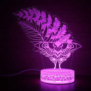 May include: A purple LED night light with a white silhouette of a butterfly and fern leaves. The light is on a round base with a textured surface.
