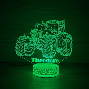 Könnte beinhalten: Grünes LED-Nachtlicht in Traktorform mit dem Namen "Theodore" in Weiß beleuchtet. Der Traktor hat große Reifen und detaillierte Merkmale. Der Sockel hat eine strukturierte, kreisförmige Form.