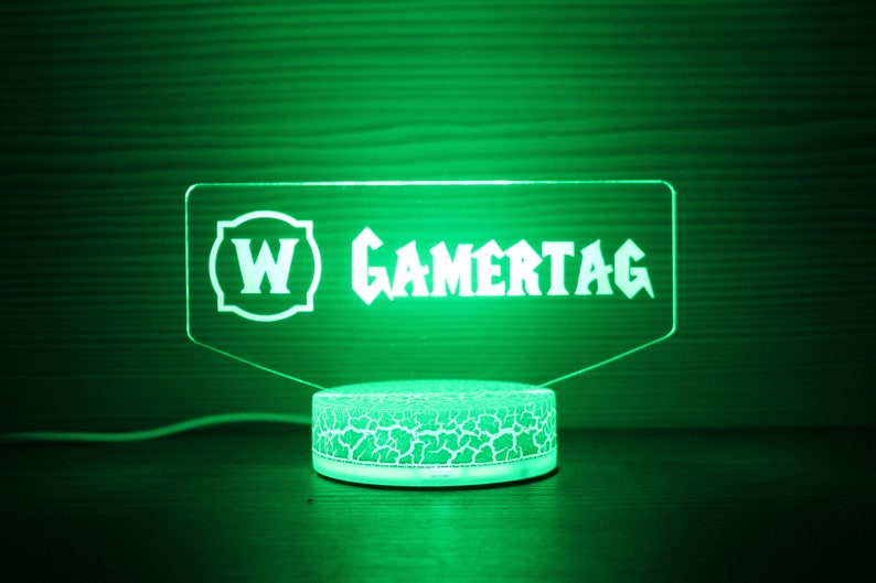 May include: A green LED light with a clear acrylic sign that reads "W GAMERTAG". The sign is mounted on a round, white base with a textured surface.