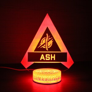 May include: A red triangular LED night light with a yellow graphic of a sword and the word "ASH" in a white rectangle. The light is on a round base with a textured design.