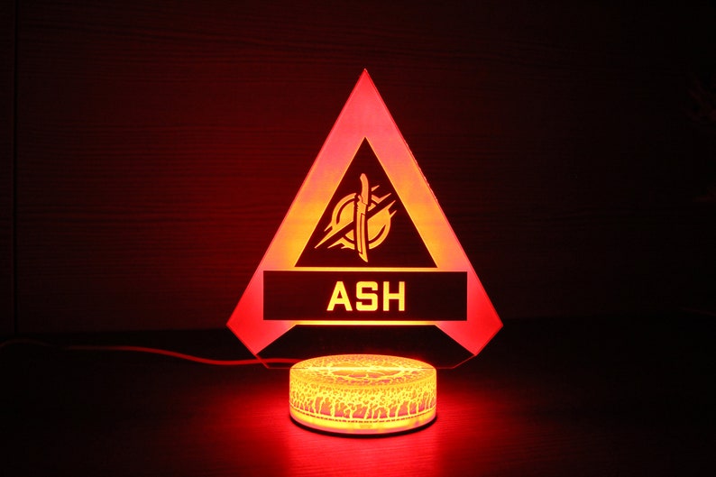 May include: A red triangular LED night light with a yellow graphic of a sword and the word "ASH" in a white rectangle. The light is on a round base with a textured design.