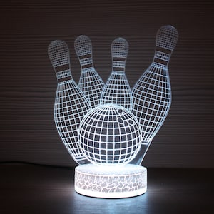 Puede incluir: Una luz nocturna impresa en 3D de color blanco con forma de bola de bolos y cinco bolos. La luz está encendida e ilumina el objeto.