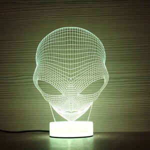 May include: A 3D-effect alien head night light. The light is white and emits a soft glow. The alien head is constructed of a grid-like pattern, and sits on a white circular base. The background is a wooden texture.