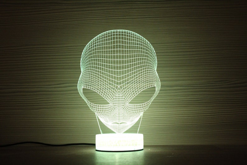 May include: A 3D-effect alien head night light. The light is white and emits a soft glow. The alien head is constructed of a grid-like pattern, and sits on a white circular base. The background is a wooden texture.
