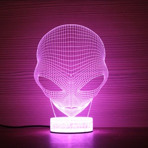 May include: A purple 3D alien head night light. The light is made of a grid of lines that form the shape of an alien's head, with large eyes and a small mouth. The base is a small, round, white light.