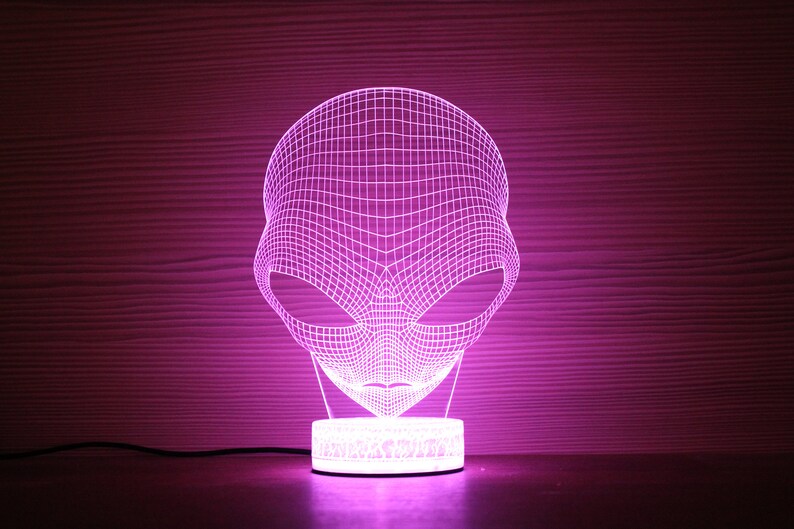 May include: A purple 3D alien head night light. The light is made of a grid of lines that form the shape of an alien's head, with large eyes and a small mouth. The base is a small, round, white light.