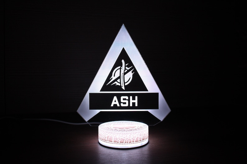 May include: A white LED light with a triangular base featuring a black and white logo with the text "ASH". The base is on a round, white, light-up platform.