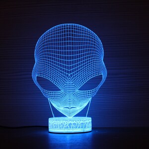 May include: A blue LED alien head lamp. The lamp has a wireframe design of an alien head, with large oval eyes. The lamp sits on a round base with a cracked pattern. The lamp emits a bright blue light.