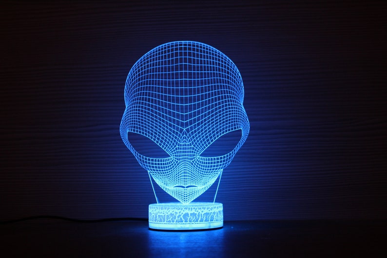 May include: A blue LED alien head lamp. The lamp has a wireframe design of an alien head, with large oval eyes. The lamp sits on a round base with a cracked pattern. The lamp emits a bright blue light.