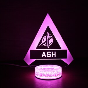 May include: A pink LED night light shaped like a triangle with the word "ASH" and a stylized logo inside. The light is on a round base with a decorative pattern.