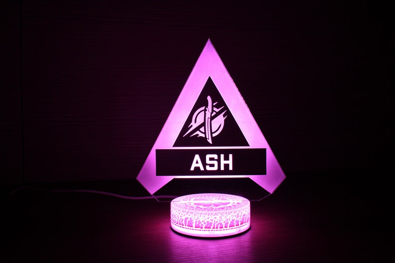 May include: A pink LED night light shaped like a triangle with the word "ASH" and a stylized logo inside. The light is on a round base with a decorative pattern.