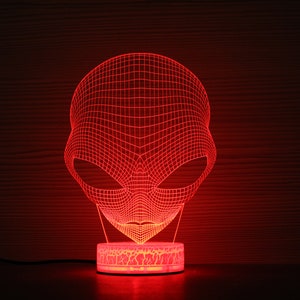 May include: A red 3D alien head night light. The alien head is made of a grid of red lines, with large oval eyes. The base of the light is a small, round, illuminated platform. The background is a dark wood grain.