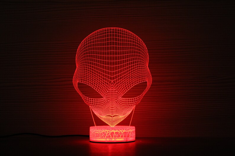 May include: A red 3D alien head night light. The alien head is made of a grid of red lines, with large oval eyes. The base of the light is a small, round, illuminated platform. The background is a dark wood grain.