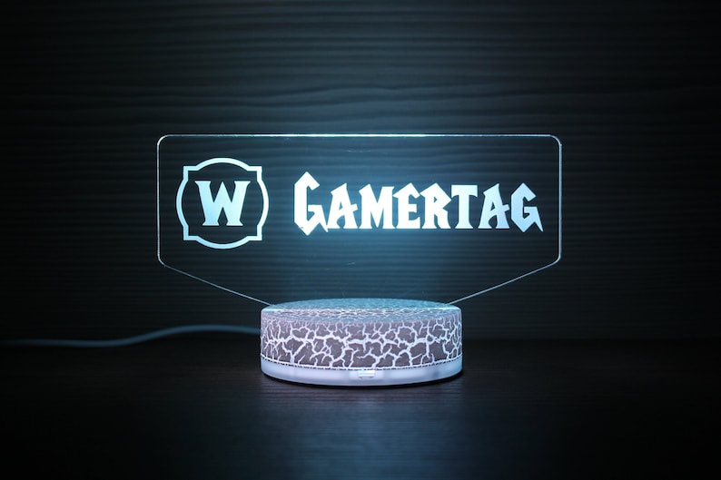 May include: A white acrylic light-up sign with the text "W GAMERTAG" in a stylized font. The sign is sitting on a white base with a textured surface.