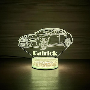 Puede incluir: Luz de noche LED con forma de coche con el nombre "Patrick" grabado debajo. El coche es un sedán, y la base de la luz tiene un patrón agrietado. La luz emite un brillo suave y cálido.