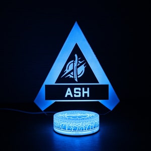 May include: A blue and white 3D printed night light shaped like a triangle with the Apex Legends character Ash's logo and name "ASH" on it.