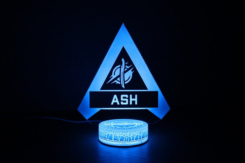 May include: A blue and white 3D printed night light shaped like a triangle with the Apex Legends character Ash's logo and name "ASH" on it.