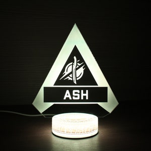 May include: A white light-up lamp shaped like a triangle with a black interior. The triangle has a white logo with a knife and the word "ASH" in white letters. The lamp is sitting on a white circular base.