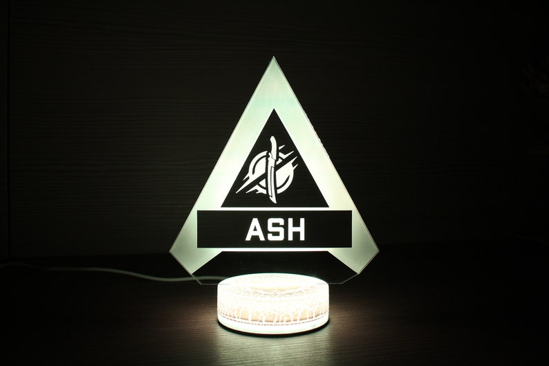 May include: A white light-up lamp shaped like a triangle with a black interior. The triangle has a white logo with a knife and the word "ASH" in white letters. The lamp is sitting on a white circular base.