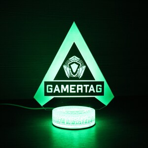 May include: A green illuminated triangle shaped lamp with a white gamertag logo and a white circular base with a geometric pattern.