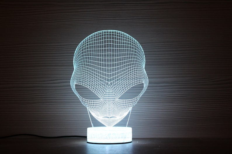 May include: A blue-glowing alien head lamp with a grid design. The lamp emits a cool blue light, creating a futuristic ambiance. The lamp sits on a round base and is connected to a power cord.